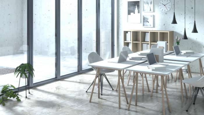 大きな窓から自然光が差し込む明るいオフィス空間。白を基調としたデスクと椅子が並び、ノートパソコンや書類が配置されている。奥には棚や壁掛け時計、観葉植物があり、シンプルで落ち着いたオフィスの雰囲気。