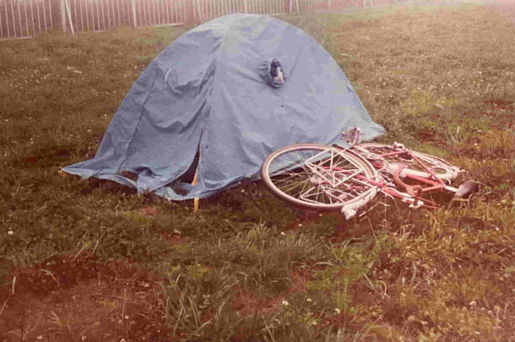 北海道・納沙布岬近くの空き地。朝霧に包まれた草の上に設営された、ソロ用の青いドーム型テント。そのすぐ横には、長旅を支えてきた赤いフレームの自転車が、倒すように置かれている。最果ての地を感じさせる、静謐でどこか寂寥感のある風景。