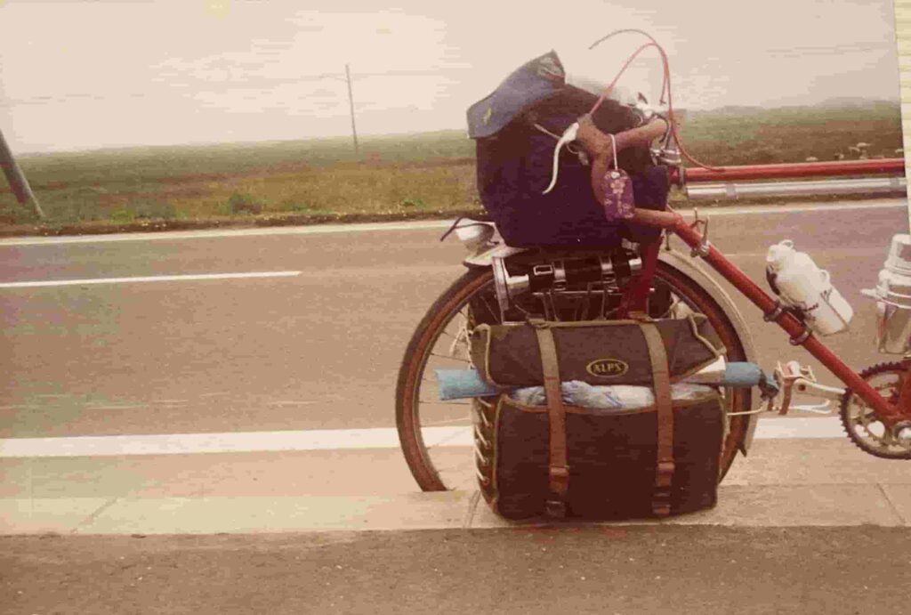 北海道のどこまでも続く直線道路の脇に停められた、赤いフレームのランドナー（旅行用自転車）。リアキャリアには大きなサイドバッグとバッグが積み上げられ、当時の長旅を物語る使い込まれた装備が印象的な、セピア色の思い出写真。