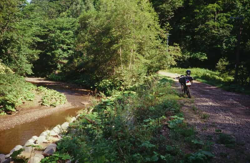 木々に囲まれた林道の脇に停められたオフロードバイクと小川