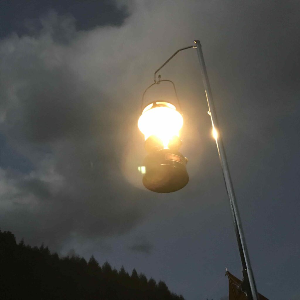 夜のキャンプ場で、ランタンスタンドに吊るされたコールマンのガソリンランタンが明るく灯っている様子。背景にはうっすらと夜空の雲と深い森のシルエットが広がっている。