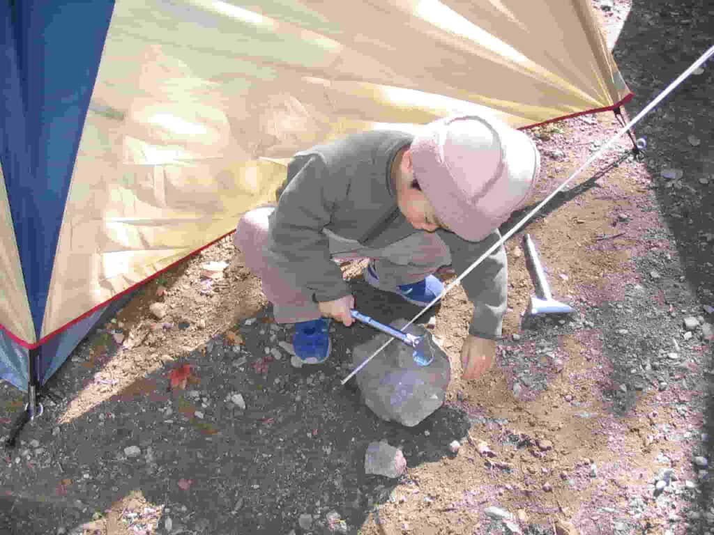 テントの脇でペグハンマーを手に石をたたく子供の姿。設営中のキャンプサイトの一場面。