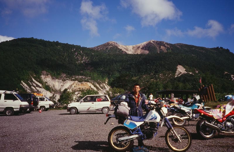 ニセコ五色温泉野営場の駐車場にて、スズキSX200とライダー。背景には、活火山特有の白い山肌を現したイワオヌプリが、青空の下にそびえ立つ。