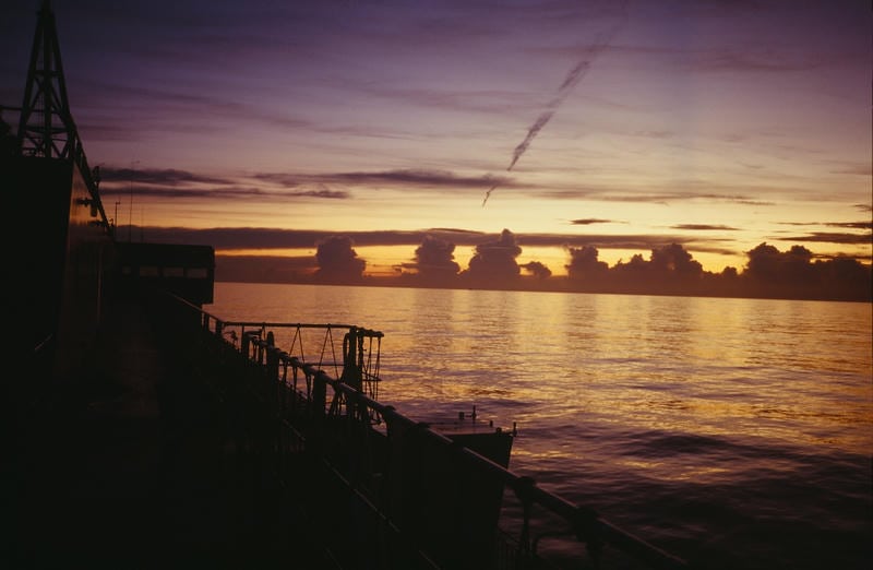 帰路のフェリーのデッキから眺める夕暮れの海。水平線には入道雲のような雲が等間隔に並び、夕日の残光が海面を黄金色に照らしている。旅の終末を感じさせる静寂な風景。
