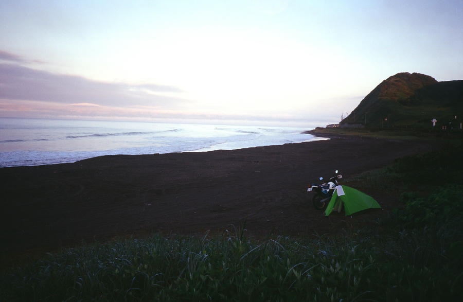 北海道の海岸線、夕暮れ時の砂浜に設営されたモンベルのムーンライト1型(グリーン)。その横には相棒のスズキSX200が停まっている。遠くに打ち寄せる波と、静かに更けゆく空が広がる情緒的なキャンプ風景。