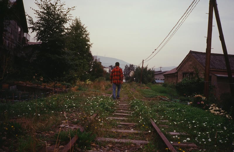 北海道・小樽の旧国鉄手宮線跡。草に覆われた廃線跡の線路の上を、赤いチェックのシャツを着た男性が遠くへ向かって歩いていく後ろ姿。