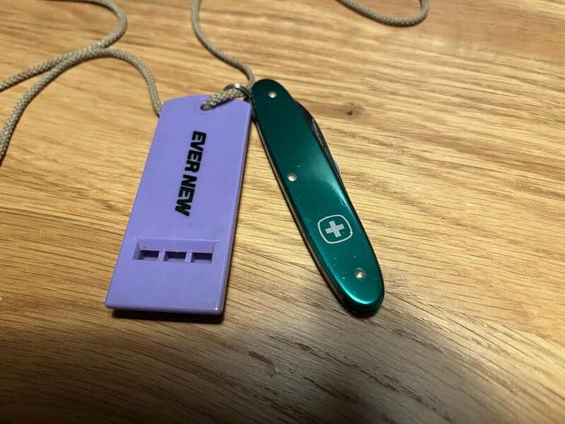 紐で繋がれたエバニュー（EVER NEW）の紫色ホイッスルと、緑色のウェンガー（Wenger）のナイフ「スーベニール」。