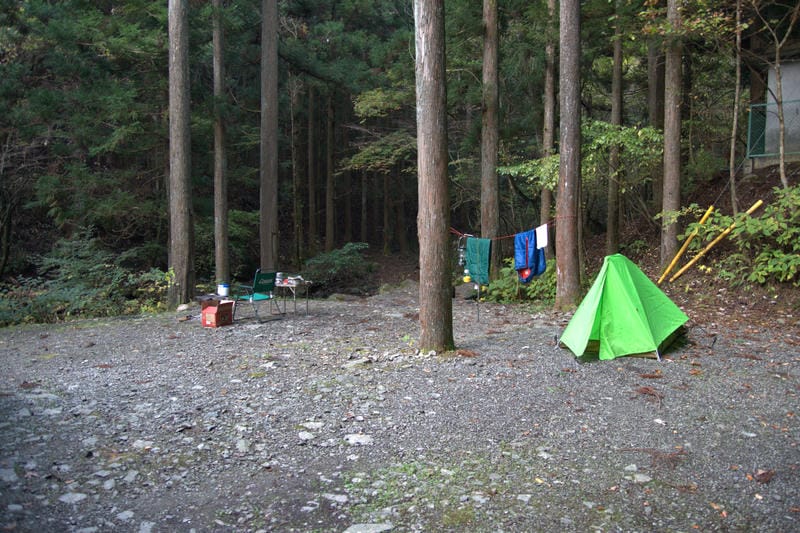 「道志の森キャンプ場」での2泊3日ソロキャンプ、2日目の昼。鮮やかな黄緑色のモンベル・ムーンライト1型テントの横、木とテントの間に張られたロープに、青と緑のシュラフ（寝袋）が干されている。テントの前には、焚き火台、チェア、テーブルが並べられたままで、ソロキャンプの生活感がにじみ出ている。