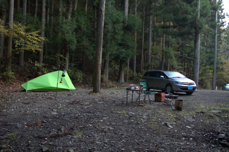 深い杉林に囲まれた山間部のキャンプ場「道志の森キャンプ場」。砂利の広場に、鮮やかな黄緑色のモンベル・ムーンライト1型テントが設営されている。テントの前にはランタンが吊るされ、その奥には車が横付けされている。手前には焚き火台、チェア、テーブルが並べられ、ソロキャンプの準備が整っている様子。