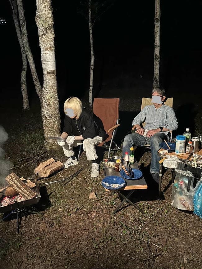 夜の白樺林のキャンプ場で、ローチェアに座って焚き火を囲みながらくつろぐ2人の人物